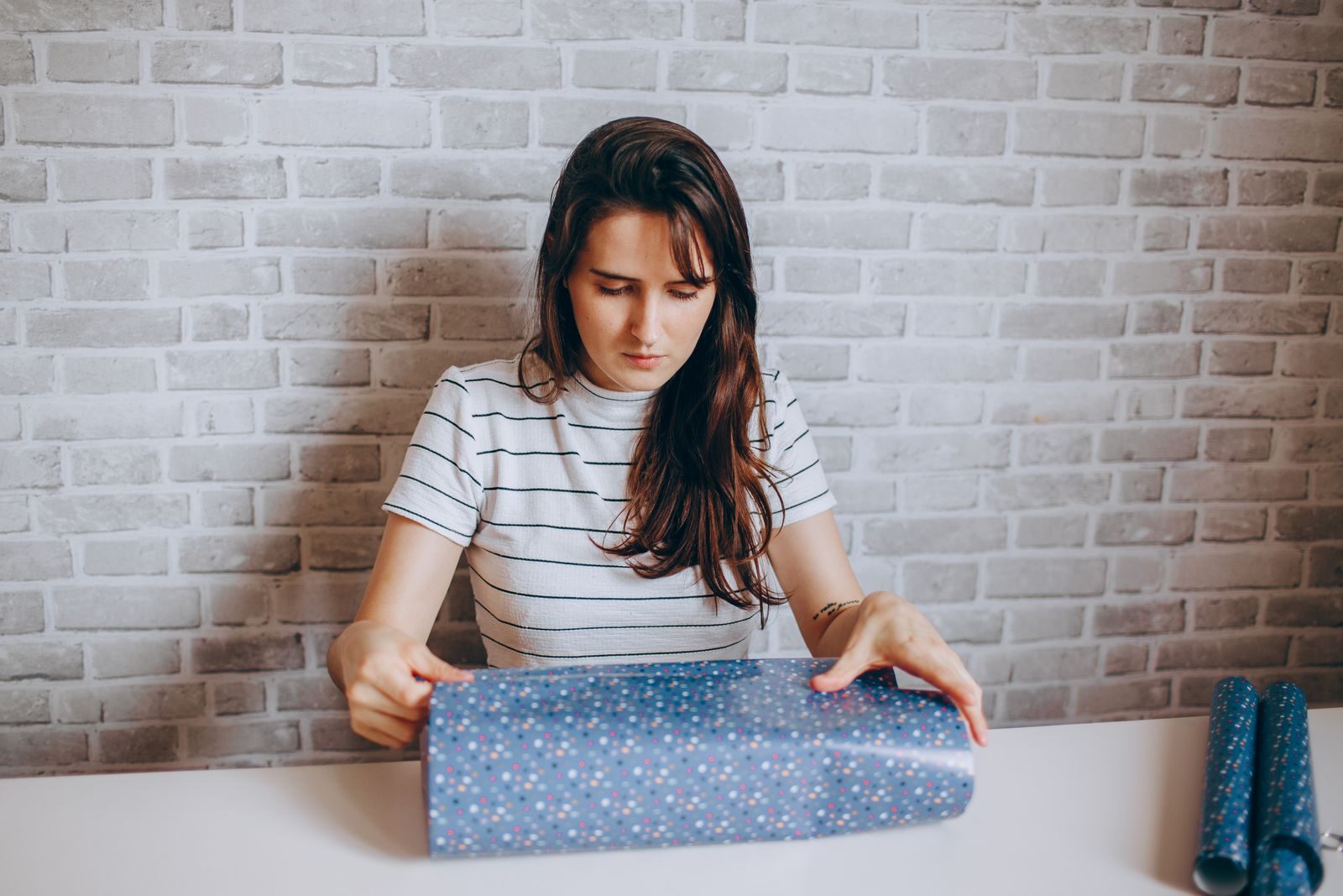 Young woman carefully wrapping a gift with decorative paper indoors against a stylish brick wall.
