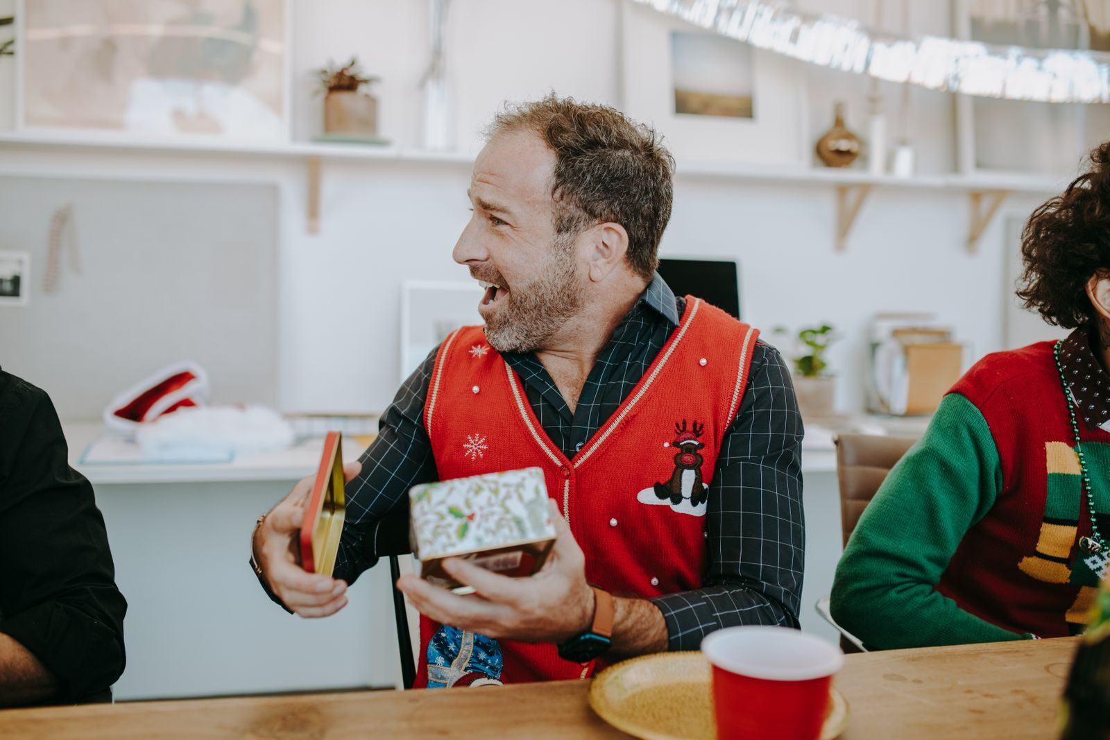 A joyful man in a festive sweater opening a gift at a Christmas home gathering.